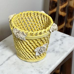 May include: A yellow ceramic basket with a woven lattice design and a rope-textured rim. The exterior is decorated with three white sculpted rose blossoms with leaves attached at intervals around the upper and middle sections of the vessel.