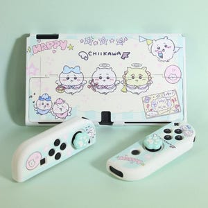 Può includere: Una console e controller Nintendo Switch azzurri e bianchi con personaggi dei cartoni animati e la parola "HAPPY". La console presenta un design decorativo con la scritta "CHIIKAWA". I controller hanno un design a impronta di zampa.