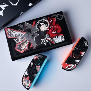 Könnte beinhalten: Schwarzes Nintendo Switch-Gehäuse und Joy-Cons mit Charakterkunst in Rot, Weiß und Grau. Das Gehäuse hat eine Grafik eines Charakters mit Maske und dem Wort "Wake Up". Die Joy-Cons sind schwarz mit roten und weißen Akzenten.