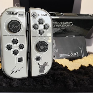 May include: Silver and black Nintendo Switch Joy-Con controllers with the text "PSTCLIC" and "FRGMT". The controllers feature black buttons, a lightning bolt design, and a Pikachu graphic. The text "BOLT PROJECT & POKÉMON" is also visible.