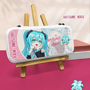 Może przedstawiać: Białe prostokątne etui z postacią anime Hatsune Miku z niebieskimi włosami i oczami. Etui ma różową obwódkę z napisem "HATSUNE MIKU". Etui jest prezentowane na małym drewnianym sztaludze na różowym tle.