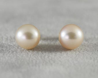 May include: Two round, lustrous, light peach-colored pearls. The pearls have a smooth surface and reflect light, creating a soft glow. The pearls are set against a neutral background, highlighting their color and shape. These are likely jewelry components.