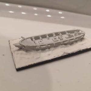 May include: A miniature gray model of a long boat with several small figures inside. The boat sits on a rectangular base with a white, textured surface, possibly representing water or ice. The boat has detailed features.