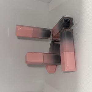 Puede incluir: Varios labiales con un dise&ntilde;o degradado. Los tubos de labial tienen una base rosa que se desvanece a un gris oscuro en la parte superior. Los labiales est&aacute;n apilados sobre un fondo blanco.