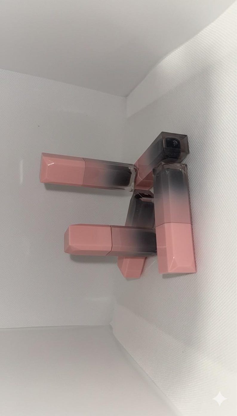 Puede incluir: Varios labiales con un dise&ntilde;o degradado. Los tubos de labial tienen una base rosa que se desvanece a un gris oscuro en la parte superior. Los labiales est&aacute;n apilados sobre un fondo blanco.