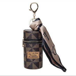 May include: A brown and tan checkered cylindrical keychain case with a gold zip and a gold keyring. A tan scarf with black trim and the letter "D" is attached. The case has a label that reads "Style And Jeans VOGUE NEW FAMOUS."