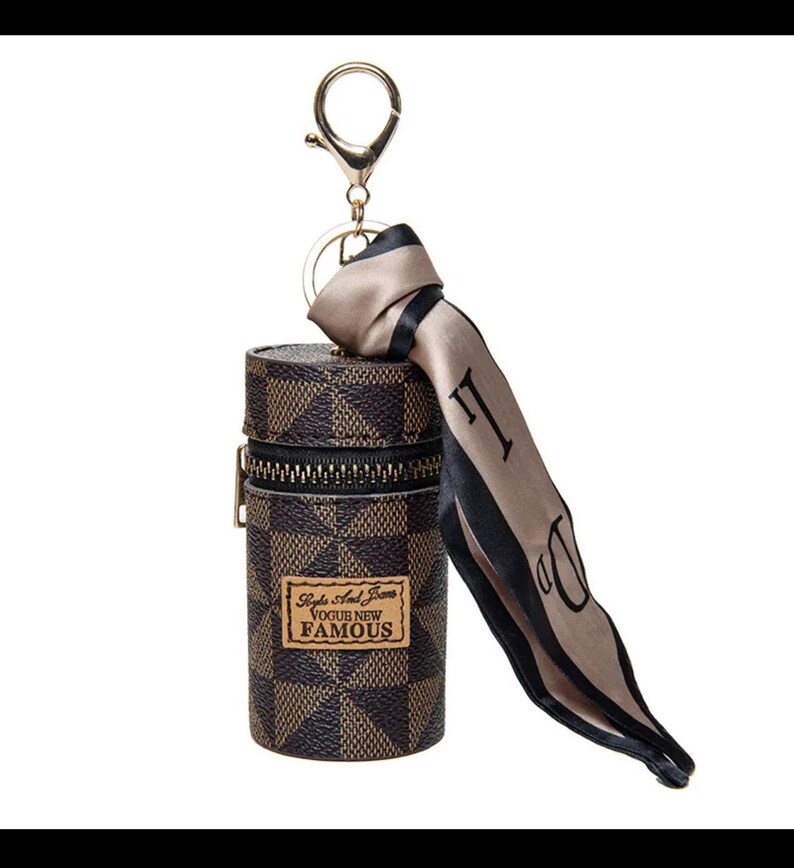 May include: A brown and tan checkered cylindrical keychain case with a gold zip and a gold keyring. A tan scarf with black trim and the letter "D" is attached. The case has a label that reads "Style And Jeans VOGUE NEW FAMOUS."