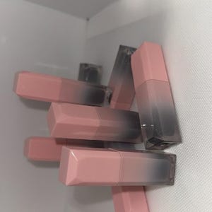 Puede incluir: Una colecci&oacute;n de barras de labios rosas y grises apiladas. Las barras de labios tienen un dise&ntilde;o geom&eacute;trico con una tapa rosa y una base gris transparente. Las barras de labios est&aacute;n en un espacio blanco texturizado.