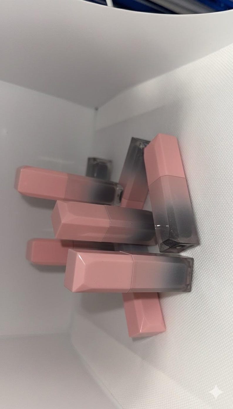 Puede incluir: Una colecci&oacute;n de barras de labios rosas y grises apiladas. Las barras de labios tienen un dise&ntilde;o geom&eacute;trico con una tapa rosa y una base gris transparente. Las barras de labios est&aacute;n en un espacio blanco texturizado.