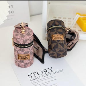 May include: Two cylindrical cases with zip closures and keyrings. One is pink with a geometric pattern, the other is brown with a similar design. Both have a label that reads "Style And Fame Vogue New Famous".