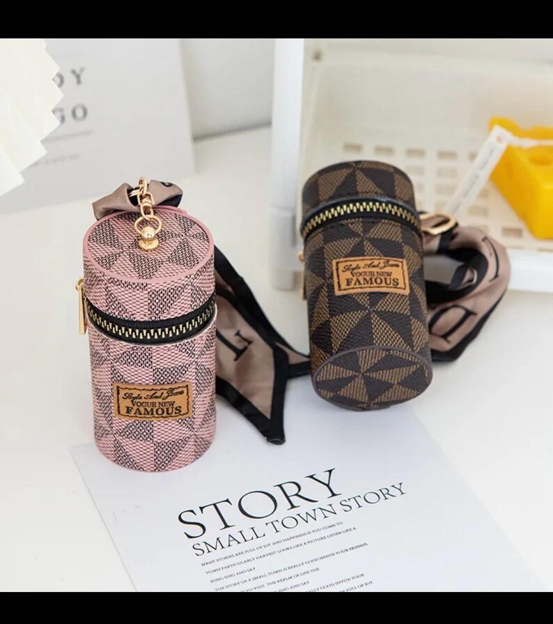 May include: Two cylindrical cases with zip closures and keyrings. One is pink with a geometric pattern, the other is brown with a similar design. Both have a label that reads "Style And Fame Vogue New Famous".
