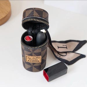 May include: A brown and tan checkered lipstick case with a zipper, containing two lipsticks. One is red, the other black. The case has a tag reading "Style And Jeans VOGUE NEW FAMOUS". A black lipstick cap is placed nearby.
