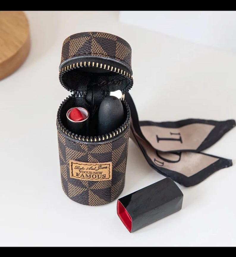 May include: A brown and tan checkered lipstick case with a zipper, containing two lipsticks. One is red, the other black. The case has a tag reading "Style And Jeans VOGUE NEW FAMOUS". A black lipstick cap is placed nearby.