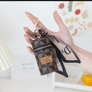 May include: A brown and black checkered pattern keychain pouch with a gold zip and a gold keyring. The pouch has a small brown label with the text "Style And Icon Vogue New Famous". A beige and black scarf is attached.