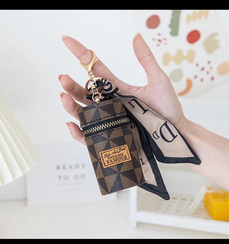 May include: A brown and black checkered pattern keychain pouch with a gold zip and a gold keyring. The pouch has a small brown label with the text "Style And Icon Vogue New Famous". A beige and black scarf is attached.