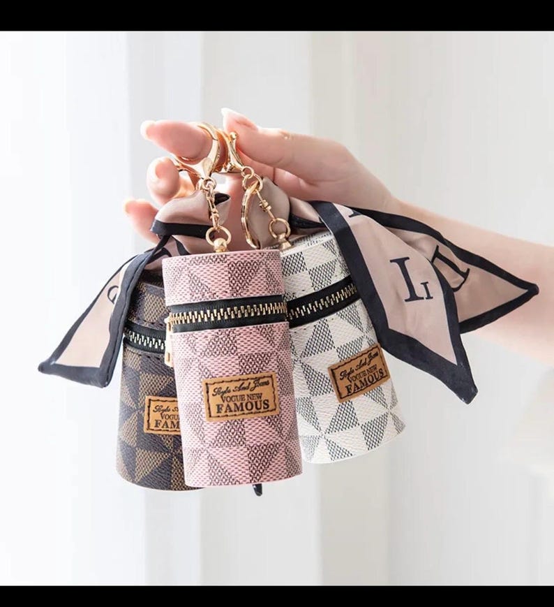 May include: Three cylindrical keyrings with gold-tone hardware and zip closures. The keyrings are patterned in brown, pink, and white with black accents. Each keyring has a small tag that reads "Style and Trend Vogue New Famous".