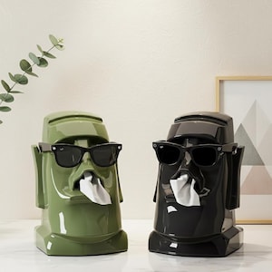 Puede incluir: Dos dispensadores de cajas de pañuelos de cerámica con forma de estatuas Moai. Uno es verde oliva, el otro es negro. Ambos llevan gafas de sol negras y tienen pañuelos blancos saliendo de la boca.