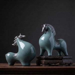 Puede incluir: Dos figuras de caballo de cerámica en un esmalte turquesa. Los caballos tienen un cuerno de unicornio y una silla de montar. Uno está sobre un soporte de madera. Las figuras están sobre una superficie de madera sobre un fondo oscuro.