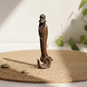 Puede incluir: Una estatua de color bronce de una figura barbuda que lleva un collar. La estatua tiene un diseño detallado y se encuentra sobre una base decorativa. La figura está de pie sobre una estera redonda y tejida con dos pequeñas piedras.