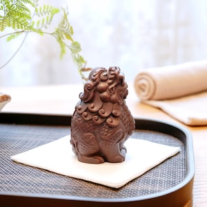 Puede incluir: Una detallada figura decorativa marrón de una criatura mítica, posiblemente un león guardián, se encuentra sobre un cuadrado blanco. La figura tiene detalles intrincados y está colocada sobre una bandeja texturizada. Una toalla enrollada y vegetación están en el fondo.