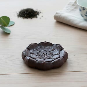 Puede incluir: Una bandeja de té marrón con forma de flor y un diseño floral detallado en el centro. La bandeja está hecha de un material oscuro y terroso y se encuentra sobre una superficie de madera clara. Hojas de té y una taza de té son visibles en el fondo.