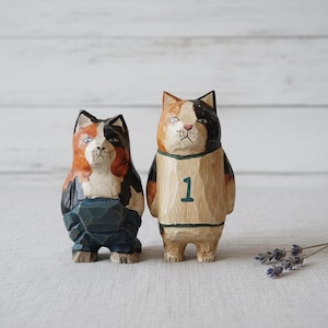 May include: Two hand-carved wooden cat figurines. One cat wears blue overalls, and the other wears a tan jersey with the number 1. Both cats have a black, orange, and white color scheme. The figurines are set against a white background.