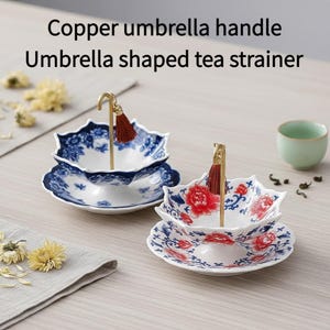 Puede incluir: Coladores de té de porcelana blanca con diseños florales azules y mangos de paraguas de cobre. Cada colador tiene un platillo a juego. Los mangos están rematados con una borla marrón. El texto de la imagen dice: "Copper umbrella handle, Umbrella shaped tea strainer."