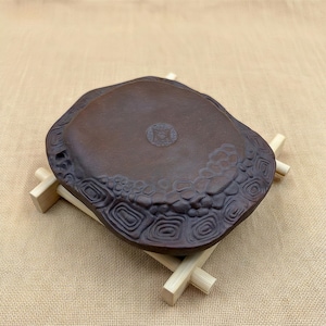 Puede incluir: Una bandeja de té de cerámica marrón, de forma irregular, con superficie texturizada y diseños decorativos. La bandeja está soportada por un soporte de madera clara, mostrando un diseño tradicional.