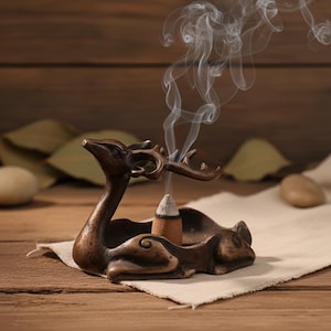 Puede incluir: Un portaincienso de color bronce en forma de ciervo. El ciervo está tumbado con la cabeza y los cuernos levantados, sosteniendo un incienso en forma de cono. Volutas de humo se elevan del incienso, creando un ambiente relajante. El soporte está sobre una tela blanca.