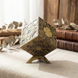 以下が含まれることがあります： 真鍮色のパズルボックス。複雑な幾何学模様と片側の円形デザインが特徴です。立方体のボックスは、お揃いのスタンドに置かれています。このボックスは、ヴィンテージでアンティークな外観をしています。