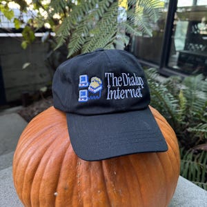 Puede incluir: Gorra de béisbol negra con el texto "The Dialup Internet" y un gráfico pixelado de una computadora. La gorra está colocada sobre una gran calabaza naranja.
