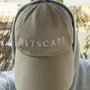 Könnte beinhalten: Khaki-farbene Baseballkappe mit dem weißen Schriftzug "NETSCAPE". Die Kappe wird mit einem grauen Kapuzenpullover getragen. Der Hut hat einen gebogenen Schirm und ein klassisches sechsteiliges Design.
