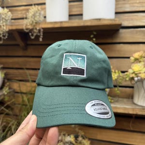 May include: A dark green adjustable baseball cap with a square patch featuring the letter "N" in a stylized design. The cap has a curved brim and a white tag that says "ADJUSTABLE".