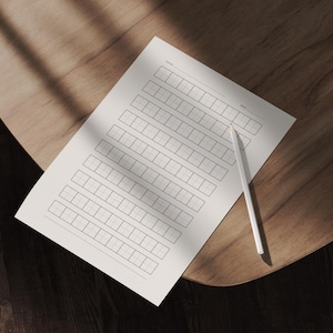 May include: A white sheet of paper with a grid of small squares for writing practice. A white pencil rests on the paper. The paper has lines for "name" and "date" at the top. The paper is on a wooden surface.