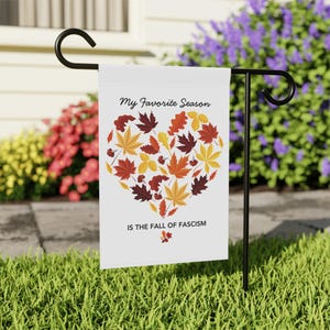 Può includere: Una bandiera da giardino bianca con la scritta "My Favorite Season" e "IS THE FALL OF FASCISM". La bandiera presenta un disegno a forma di cuore fatto di foglie autunnali nei toni del rosso, arancione e giallo. La bandiera è esposta su un supporto in metallo nero.