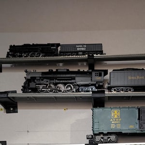 May include: A collection of model trains displayed on a wall. The trains are various sizes and colors, including black locomotives and green freight cars. Some cars have text like "Santa Fe" and "Chief West". The display includes multiple shelves.