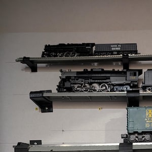 May include: A collection of model trains displayed on a wall. The trains are various sizes and colors, including black locomotives and green freight cars. Some cars have text like "Santa Fe" and "Chief West". The display includes multiple shelves.