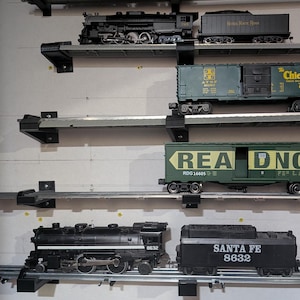 May include: A collection of model trains displayed on a wall. The trains are various sizes and colors, including black locomotives and green freight cars. Some cars have text like "Santa Fe" and "Chief West". The display includes multiple shelves.