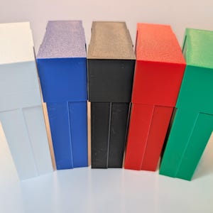 May include: Five rectangular storage cases standing upright in a row. The cases are colored white, blue, black, red, and green. Each case features a flat top lid and a vertical body with a smooth, matte finish, arranged side by side in a line.