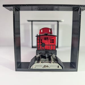 May include: A red miniature caboose model with black detailing is positioned on a section of railway track. It's displayed inside a black, rectangular frame, offering a distinctive presentation. A similar, smaller frame is in the background.