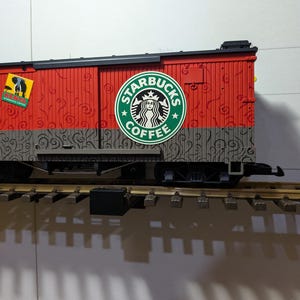 May include: A model train car with a red upper section and gray lower section. The car features Starbucks Coffee branding and other circular stickers. The car is on a model train track.