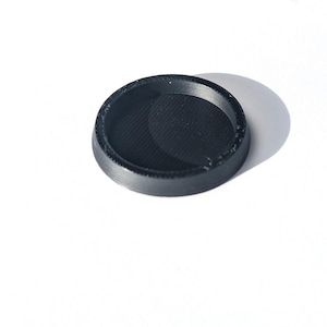 May include: A small, circular, black plastic component with a raised outer rim and a recessed, textured center. The object features a distinct layered finish consistent with 3D printing, showing a flat base and a slightly angled, circular top edge.