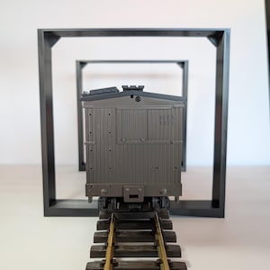 May include: A grey model train carriage on a track, framed by three black square structures. The carriage features detailed paneling and a roof. The track is made of brown and gold materials, creating a sense of depth.
