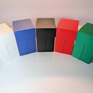 May include: Five rectangular storage boxes arranged in a row, featuring a solid matte finish in white, blue, black, red, and green. Each box has a vertical rectangular panel on the front face and a flat top surface, constructed from a rigid material.