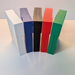 May include: Five rectangular cases arranged in a fan shape, each featuring a distinct solid color. The set includes one white, one blue, one black, one red, and one green case, all standing upright with a smooth, matte finish and clean edges.