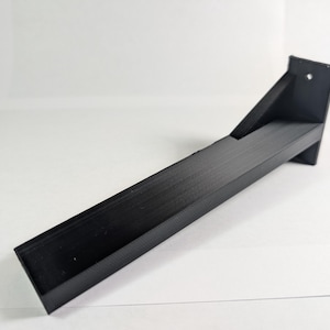May include: A black, angled shelf bracket with a rectangular base and a supporting angled piece. The bracket has a hole near the top for mounting. The surface has a textured, ridged pattern.