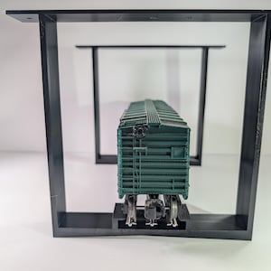 May include: A dark green model train carriage is positioned on a black metal frame. The frame is rectangular with angled legs. The carriage has detailed features and is centrally placed within the frame.
