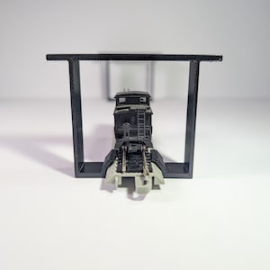 May include: A black model train locomotive on a track, set within a black rectangular frame. The locomotive has detailed features such as a ladder and mechanical parts. The scene is set against a plain white background.