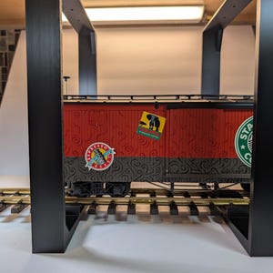 May include: A model train car featuring a red and black body with swirling patterns, positioned on gold-colored tracks. The side displays a Starbucks Coffee logo, a circular Espresso Roast emblem, and a rectangular sticker depicting an elephant.