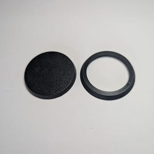 May include: A black circular plastic base with a textured top surface and a separate black circular ring with a beveled edge. Both items are solid black and feature a matte finish, designed as components for miniature figures or gaming models.